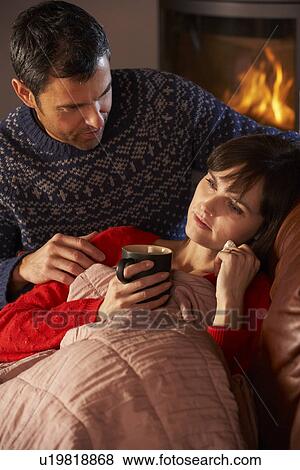Stock Photo - Husband Nursing Sick Wife With Cold Resting On Sofa By Cosy Log Fire. Fotosearch