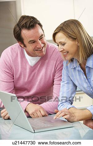 Stock Image - Mature couple using laptop in domestic kitchen. Fotosearch