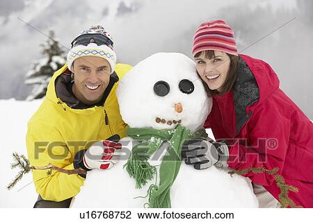 Stock Image - Middle Aged Couple Building Snowman On Ski Holiday In Mountains. Fotosearch