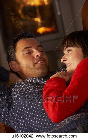 Stock Image - Middle Aged Couple Chatting On Sofa By Cosy Log Fire. Fotosearch