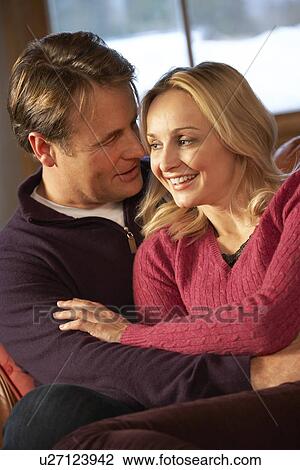 Stock Image - Middle Aged Couple Chatting On Sofa In Chalet With Winter View. Fotosearch