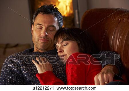 Stock Image - Middle Aged Couple Cuddling On Sofa By Cosy Log Fire. Fotosearch