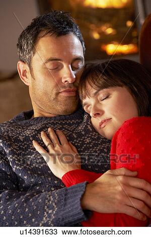 Stock Image - Middle Aged Couple Cuddling On Sofa By Cosy Log Fire. Fotosearch