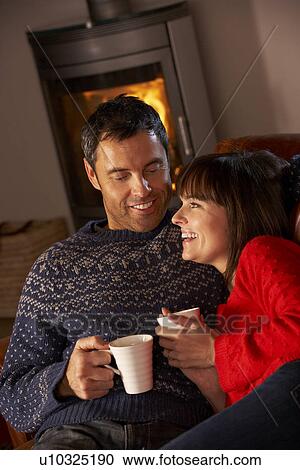 Stock Image - Middle Aged Couple Sitting On Sofa By Cosy Log Fire With Hot Drink. Fotosearch