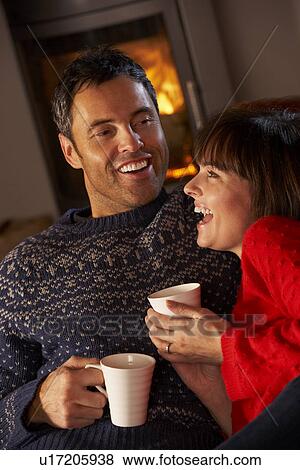 Stock Photo - Middle Aged Couple Sitting On Sofa By Cosy Log Fire With Hot Drink. Fotosearch