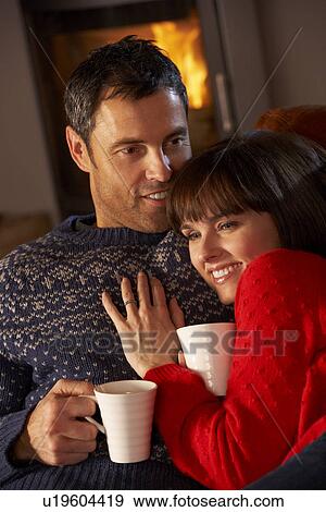 Stock Photo - Middle Aged Couple Sitting On Sofa By Cosy Log Fire With Hot Drink. Fotosearch