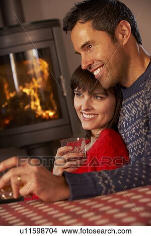 Picture - Middle Aged Couple Sitting On Sofa Watching TV By Cosy Log Fire. Fotosearch