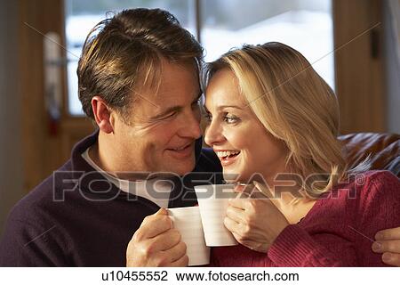 Stock Image - Middle Aged Couple Sitting On Sofa With Hot Drinks. Fotosearch