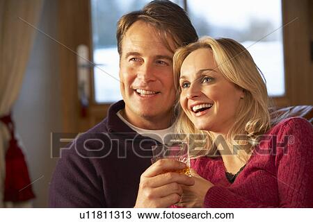 Stock Image - Middle Aged Couple Sitting On Sofa With Glasses Of Whisky Watching TV. Fotosearch