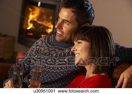 Stock Image - Middle Aged Couple Sitting Sofa By Cosy Log Fire With Glass Of Champagne. Fotosearch