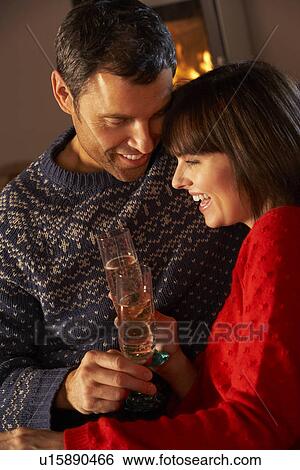 Stock Photograph - Middle Aged Couple Sitting Sofa By Cosy Log Fire With Glass Of Champagne. Fotosearch