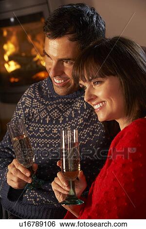 Stock Photograph - Middle Aged Couple Sitting Sofa By Cosy Log Fire With Glass Of Champagne. Fotosearch