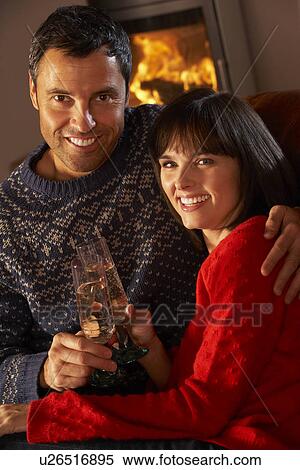 Stock Photography - Middle Aged Couple Sitting Sofa By Cosy Log Fire With Glass Of Champagne. Fotosearch