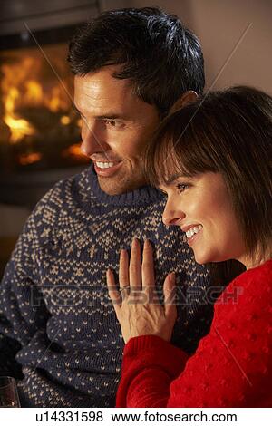 Stock Photo - Middle Aged Couple Sitting Sofa Watching TV By Cosy Log Fire. Fotosearch