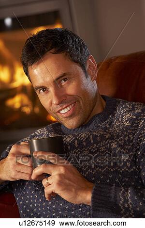 Stock Photo - Middle Aged Man Relaxing With Hot Drink By Cosy Log Fire. Fotosearch