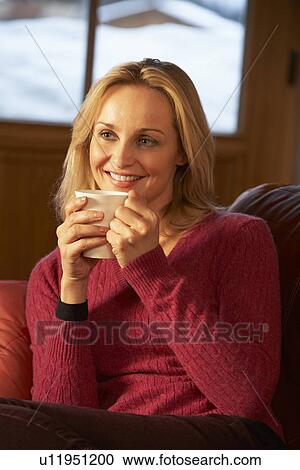 Stock Image - Middle Aged Woman Relaxing With Hot Drink On Sofa Watching TV. Fotosearch