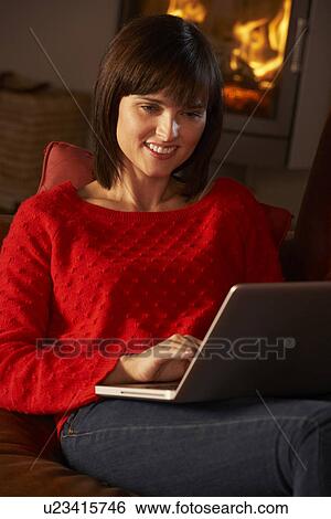 Stock Photograph - Middle Aged Woman Using Laptop Computer By Cosy Log Fire. Fotosearch