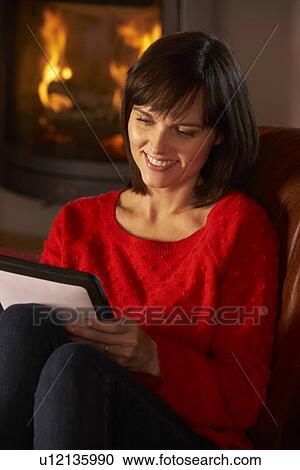 Stock Image - Middle Aged Woman Using Tablet Computer By Cosy Log Fire. Fotosearch