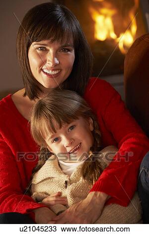 Stock Image - Mother And Daughter Relaxing On Sofa By Cosy Log Fire. Fotosearch