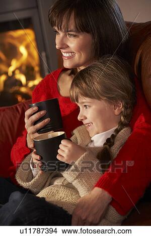 Picture - Mother And Daughter Relaxing With Hot Drink Watching TV By Cosy Log Fire. Fotosearch