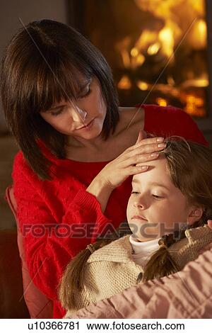 Stock Image - Mother Comforting Sick Daughter On Sofa By Cosy Log Fire. Fotosearch