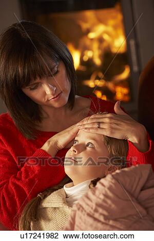 Stock Image - Mother Comforting Sick Daughter On Sofa By Cosy Log Fire. Fotosearch