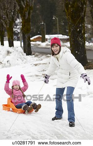 Stock Image - Mother Pulling Daughter On Sledge Along Snowy Street In Ski Resort. Fotosearch