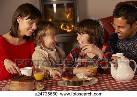 Stock Image - Portrait Of Family Enjoying Tea And Cake By Cosy Log Fire. Fotosearch