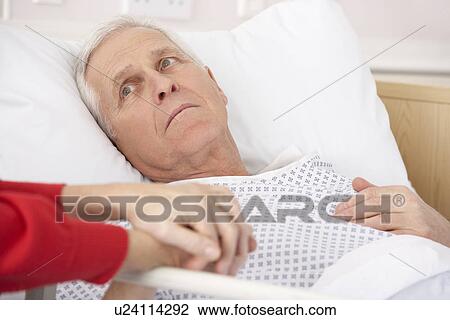 Stock Image - Senior man in hospital bed holding wife's hand. Fotosearch