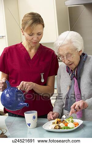 Stock Image - Senior woman with carer eating meal at home. Fotosearch