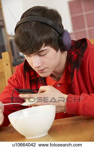 Stock Image - Teenage Boy Listening To MP3 Player Whilst Eating Breakfast. Fotosearch