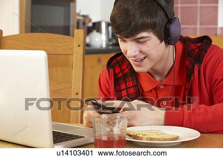 Stock Image - Teenage Boy Using Laptop And Listening To MP3 Player Whilst Eating Breakfast. Fotosearch