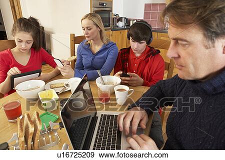 Stock Photo - Teenage Family Using Gadgets Whilst Eating Breakfast Together In Kitchen. Fotosearch
