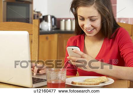 Stock Photo - Teenage Girl Using Laptop And Mobile Phone Whilst Eating Breakfast. Fotosearch