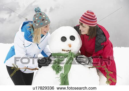 Stock Photograph - Two Female Friends Building Snowman On Ski Holiday In Mountains. Fotosearch