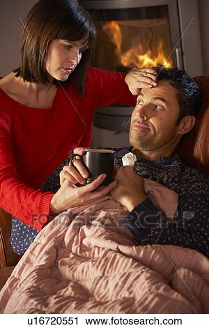 Stock Image - Wife Nursing Sick Husband With Cold Resting On Sofa By Cosy Log Fire. Fotosearch