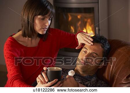 Stock Photograph - Wife Nursing Sick Husband With Cold Resting On Sofa By Cosy Log Fire. Fotosearch