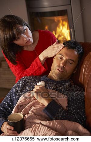 Stock Photography - Wife Nursing Sick Husband With Cold Resting On Sofa By Cosy Log Fire. Fotosearch