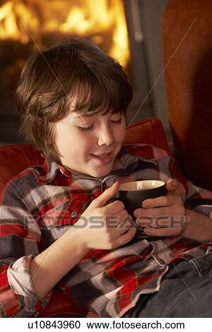 Stock Image - Young Boy Relaxing With Hot Drink By Cosy Log Fire. Fotosearch