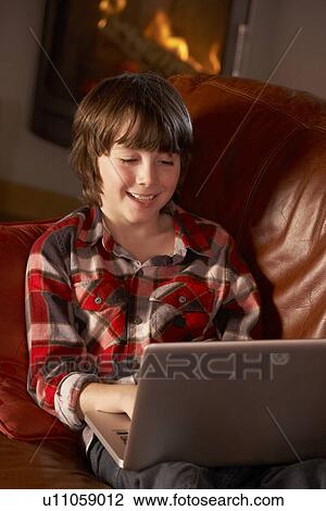Stock Image - Young Boy Relaxing With Laptop By Cosy Log Fire. Fotosearch