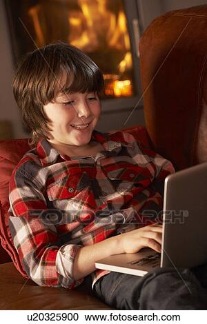 Stock Image - Young Boy Relaxing With Laptop By Cosy Log Fire. Fotosearch