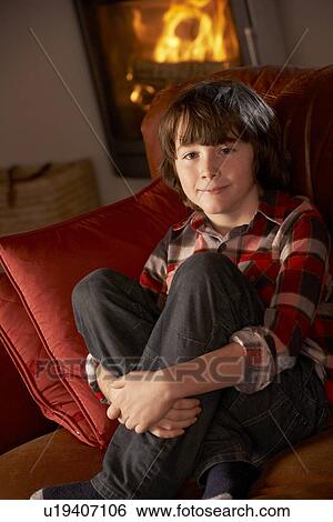 Stock Photograph - Young Boy Sitting On Sofa By Cosy Log Fire. Fotosearch