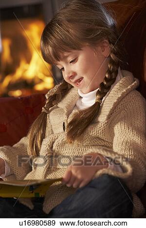 Stock Photo - Young Girl Sitting On Sofa And Reading Book By Cosy Log Fire. Fotosearch