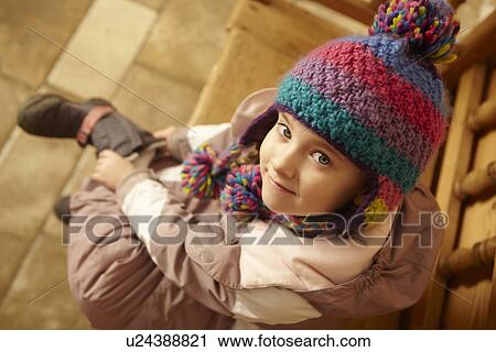 Stock Image - Young Girl Sitting On Wooden Seat Putting On Warm Outdoor Clothes And Boots. Fotosearch