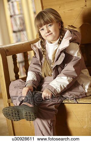 Stock Image - Young Girl Sitting On Wooden Seat Putting On Warm Outdoor Clothes And Boots. Fotosearch
