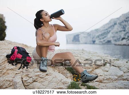 Stock Photo - France, Marseille, Woman with backpack drinking water on rocky beach. Fotosearch