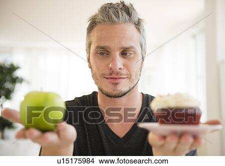 Man weighing green apple against cup cake View Large Photo Image Picture - Man weighing green apple against cup cake. Fotosearch