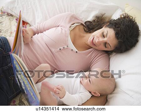 Picture - Mother and baby boy (2-5 months) lying together on bed, reading book. Fotosearch
