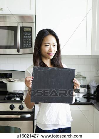 Stock Photo - Portrait of smiling young woman holding blackboard in kitchen. Fotosearch