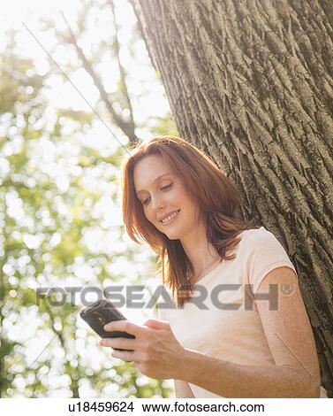 Picture - Woman leaning at tree and texting. Fotosearch